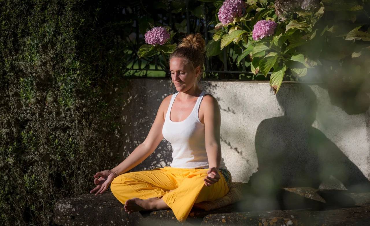 Patricia sentada em meditação num banco de pedra da Quinta do Pé Descalço, em Sintra, com hortênsias em flor atrás.