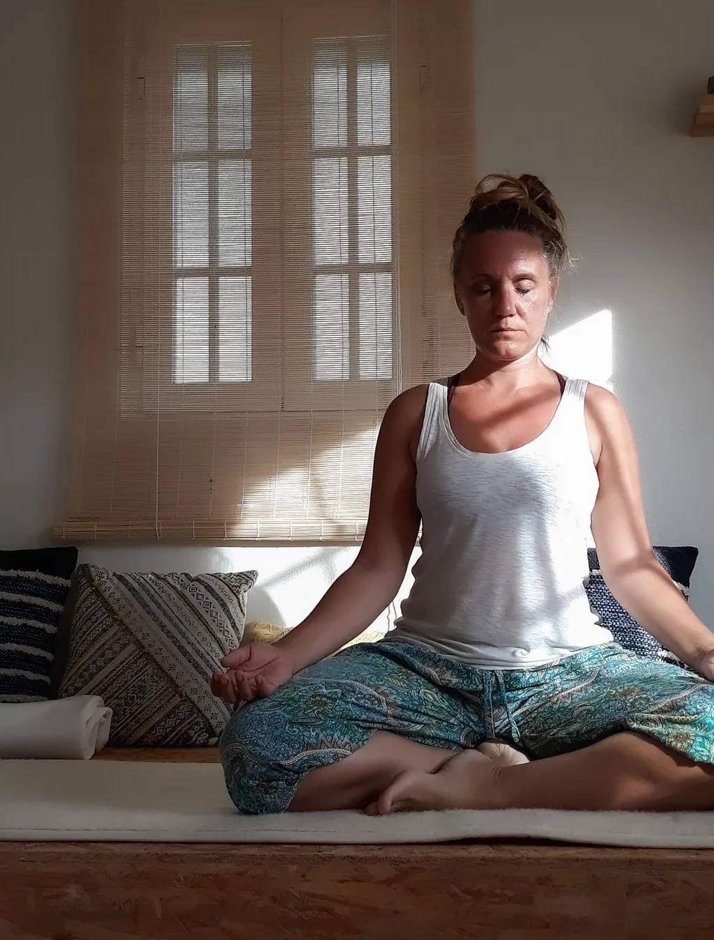 Patricia sentada em meditação na sala, de olhos fechados e com um mala na mão, à luz da janela.